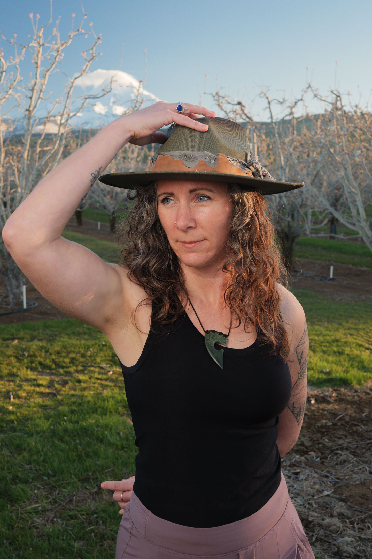 Kate Ensor holding her hat brim in the orchard with mountain behind her